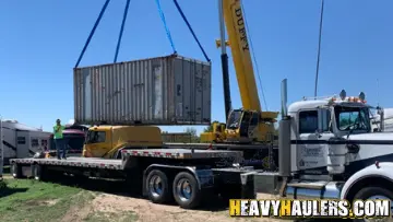 shipping container being loaded by cranes onto a step deck trailer